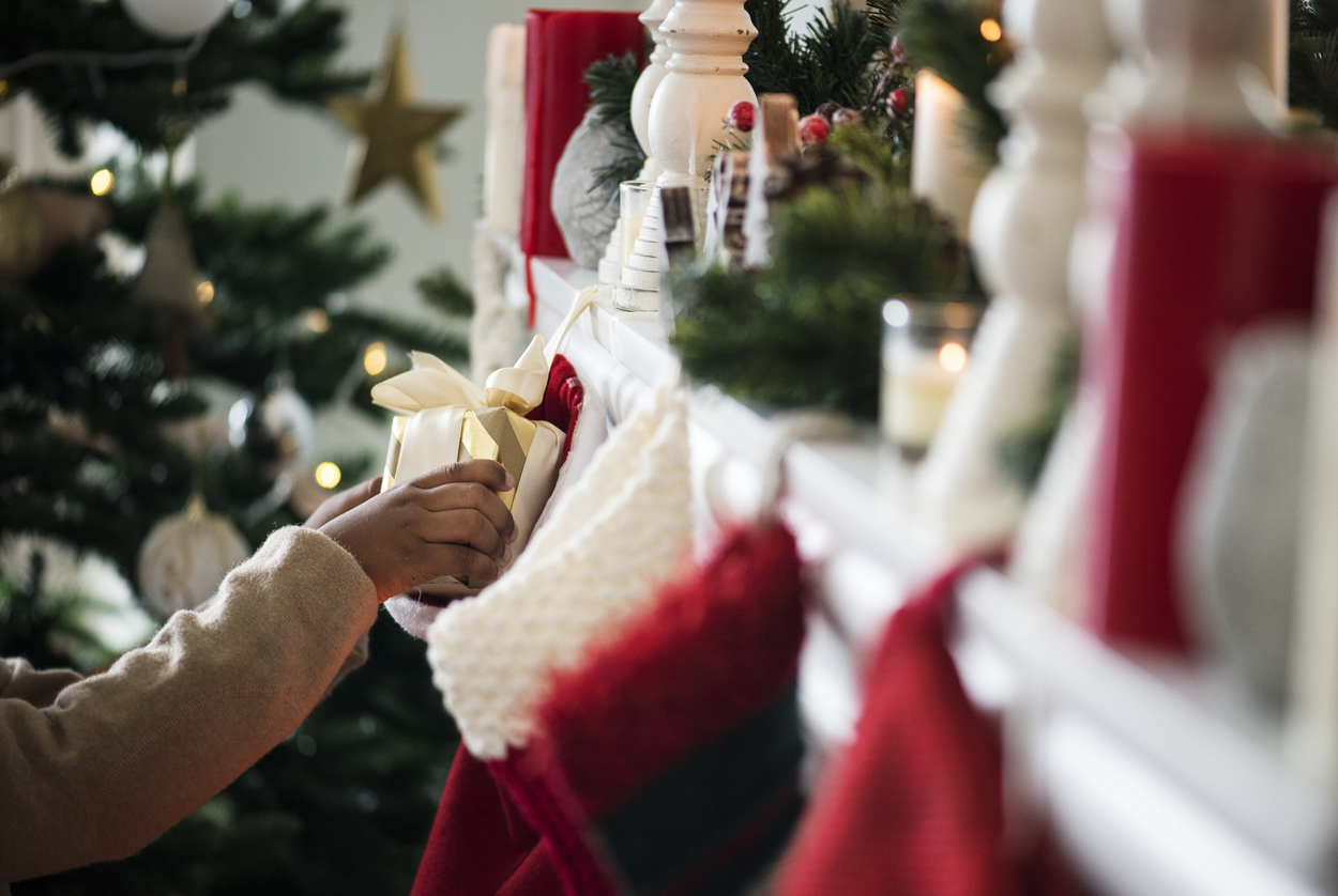 Christmas stockings hang off a mantle in front of a tree. A small child's arms are seen reaching into one of the stockings and pulling out a gift. Lotteries Warn: Do Not Buy Kids Lottery Tickets for the Holidays