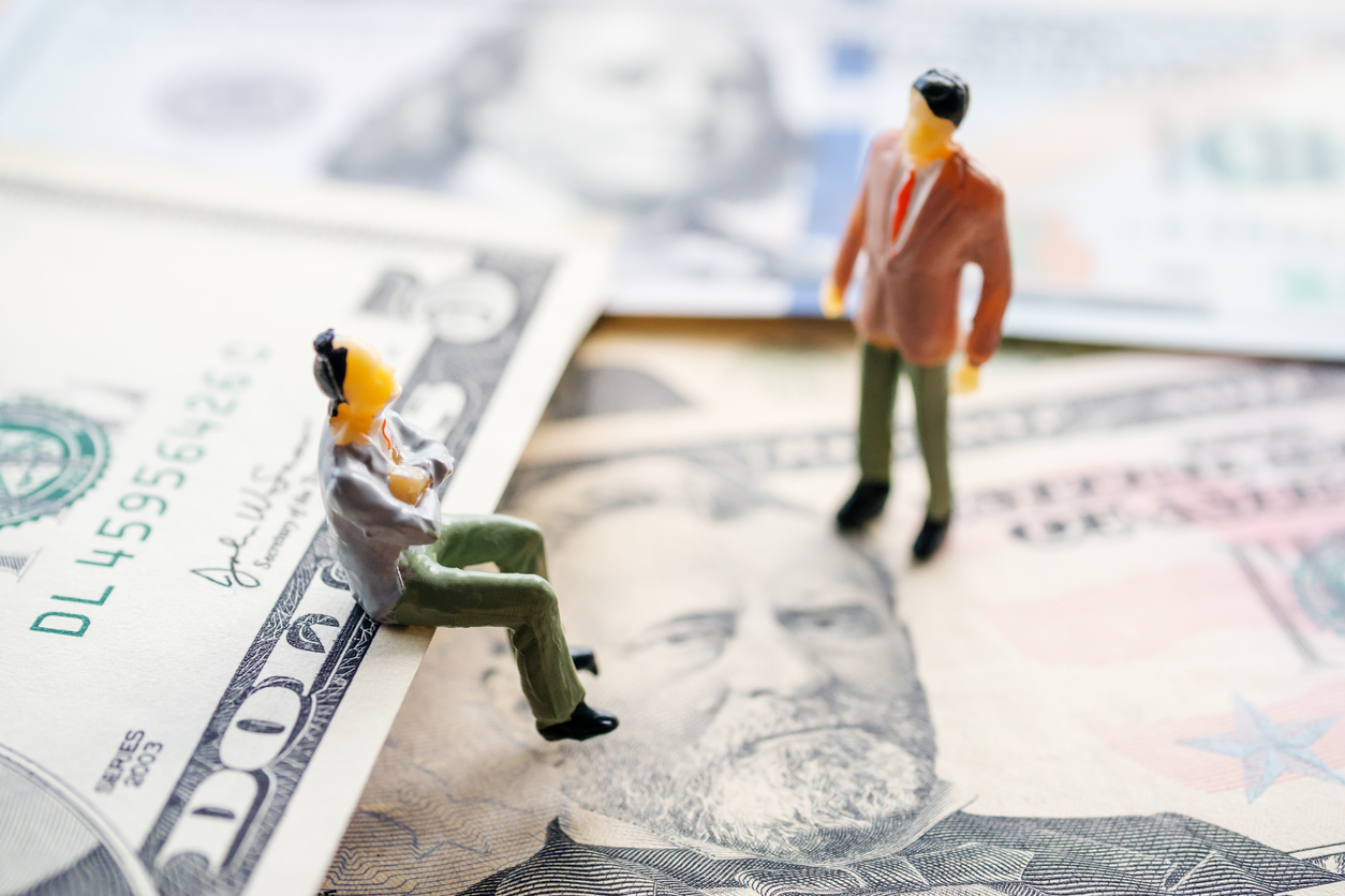 Two tiny toy figures of men in suits -- one standing and one sitting with his arms crossed on the edge of a 5 dollar bill. other bills are spread out beneath and around them.